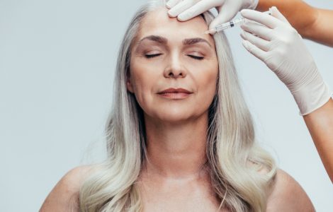 Doctor with gloves injecting a botox with an needle into temple of the senior woman against grey background. Woman getting anti aging injection on her face to reduce wrinkles.